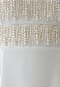 Bílá látka s vrstveným designem, která na vrchu obsahuje kaskádovité prameny perel a korálků, čímž vytváří texturovaný dekorativní prvek.