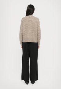Frau, die sich mit dem Rücken zur Kamera dreht, trägt einen beigen Strickpullover und weit geschnittene schwarze Hosen vor einem schlichten weißen Hintergrund.