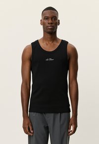 Μαύρο ριμπ φανελάκι με στρογγυλή λαιμόκοψη και κεντημένο λογότυπο στο στήθος· φοριέται με γκρι παντελόνι.