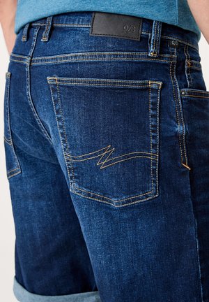 Pantalones cortos de mezclilla azul oscuro con dobladillo enrollado, costuras amarillas prominentes y dos bolsillos traseros con un detalle de diseño en zigzag.