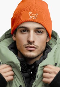 Orange knit beanie with a dog silhouette patch, worn with a green parka. Contains ribbed texture and a close-fitting design.