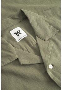 Chemise à manches courtes vert olive avec un col, dotée d'un bouton blanc et d'une étiquette avec le logo "W.W" brodé à l'intérieur. Texture de tissu lisse.