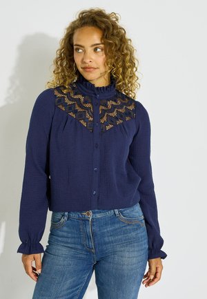 Mujer con cabello rizado que lleva una blusa azul marino con detalle de encaje en el pecho y mangas largas, combinada con vaqueros azules.