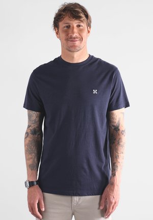 Homme avec des tatouages sur les deux bras portant un t-shirt bleu marine et un pantalon beige, se tenant devant un fond clair et uni avec un léger sourire.