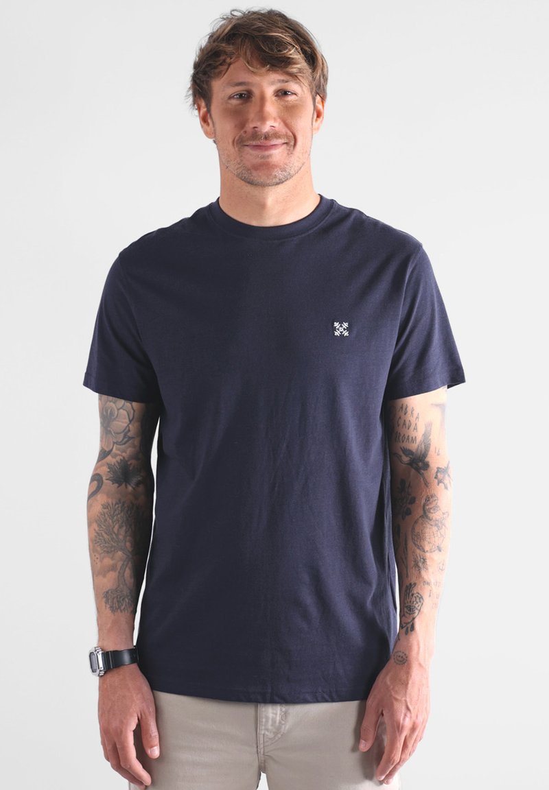 Homme avec des tatouages sur les deux bras portant un t-shirt bleu marine et un pantalon beige, se tenant devant un fond clair et uni avec un léger sourire.