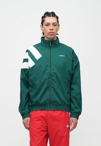 Mladý muž s vrkočmi, oblečený v zelenej vetrovke Adidas so zipsom a bielymi pruhmi a červených športových nohaviciach Adidas, stojaci pred jednoduchým pozadím.