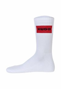 Weiße Baumwollmischsocken mit gerippter Textur, die ein rotes rechteckiges Band mit dem schwarzen Wort "HUGO" am Bund aufweisen.