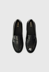 Chaussures noires en cuir à enfiler, avec un accent en verni brillant sur les orteils et une texture semblable au daim sur les côtés. Comprend un logo doré.