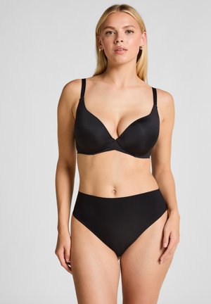 Mujer con cabello rubio largo que lleva un sujetador negro con relleno y ropa interior negra de talle alto, de pie contra un fondo claro y liso.