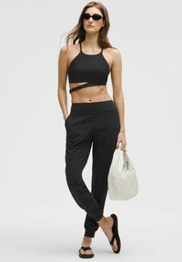 Schwarzes, kurzes Halstops mit Wickeldetails, schwarze, schmal zulaufende Hosen und schwarze Flip-Flops. Das Model trägt eine weiße Handtasche. Glatte Stoffoberfläche.