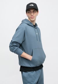 Νεαρός άνδρας με μπλε φούτερ με κουκούλα και ταιριαστό παντελόνι, με τα χέρια στις τσέπες, κοιτάζοντας πλάγια, και μαύρο καπέλο The North Face.