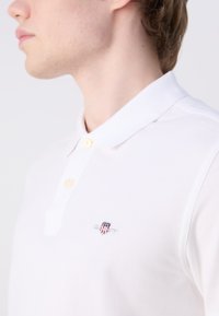 Young man wearing a white polo shirt with a small red, white, and blue shield logo on the left chest, looking to the side.
