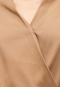 Εγχώριο πλάνο ενός ατόμου που φοράει μπεζ ρούχο με V-λαιμόκοψη τύπου wrap, με λείο, ματ ύφασμα και λεπτές υφές.