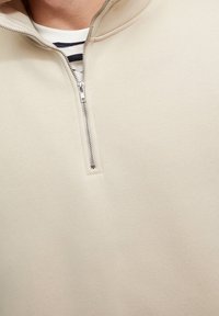 Beige lynlåsjakke med høj krave, udstyret med en metallisk lynlås og glat, struktureret stof. Minimalistisk design uden mønstre.