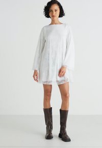 Rochie albă cu mâneci lungi și textură din dantelă, cu o croială dreaptă și decolteu rotund. Pur tată cu cizme maro până la genunchi.