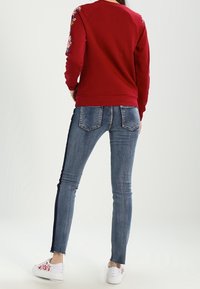 Sudadera roja con bordado floral en las mangas, combinada con vaqueros pitillo azules. Zapatillas blancas con detalles florales completan el conjunto.