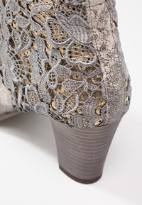 Chaussure à talons hauts grise avec une surface texturée. Comprend une dentelle finement brodée avec des accents dorés et un talon lisse et orné de motifs.