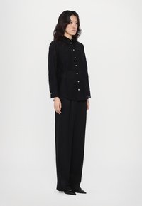 Zwarte button-up shirt, semi-doorzichtig materiaal, lange mouwen, reguliere kraag, gecombineerd met losse zwarte broeken en puntige zwarte schoenen.