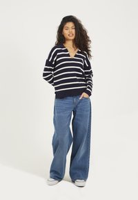 Maglione a righe navy e bianche con scollo a V, abbinato a jeans in denim blu dal taglio ampio e scarpe da ginnastica bianche.
