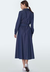 Sukienka koszulowa z denimu, ciemnoniebieska, długie rękawy, mankiety na guziki, wcięcie w talii z pasującym paskiem, do kostek, noszona z białymi trampkami.