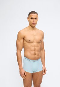 Ευθυτενής άνδρας με κοντά μαλλιά, φοράει ανοιχτό μπλε εσώρουχα Calvin Klein boxer, στέκεται σε χαλαρή στάση μπροστά σε απλό ανοιχτό φόντο.