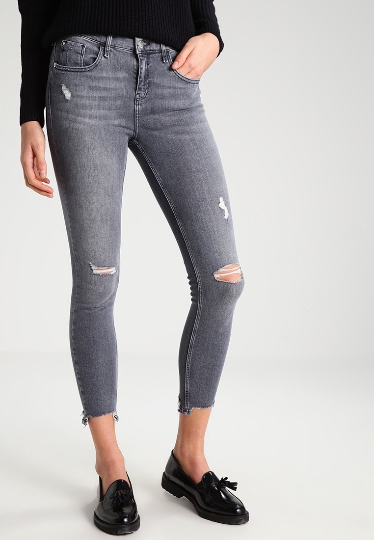 Vaqueros skinny grises en un diseño desgastado con rasgaduras en las rodillas y dobladillos deshilachados. Hechos de mezclilla, presentan un estilo clásico de cinco bolsillos.