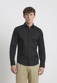 Schwarzes Langarmhemd mit Button-Down-Kragen, das eine glatte Textur und robuste Knöpfe aufweist, kombiniert mit beigen Hosen.