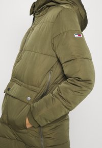 Person wearing a green padded hooded jacket with zipped side pocket and snap-button flap pocket, hand inside front pocket, small logo on sleeve.