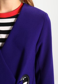 Detail osoby v fialovém saku přes pruhovanou černobílou košili s červeným límcem na světlém pozadí.