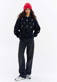 Černá mikina s kapucí s květinovou výšivkou, volný střih, klokánká kapsa, v kombinaci s černými džíny s vysokým pasem a šedými sportovními botami. Červená pletená čepice.