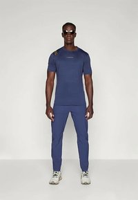 Tenue de sport bleu marine comprenant un t-shirt à manches courtes avec des rayures texturées et un logo, associé à un pantalon slim assorti et des baskets blanches.