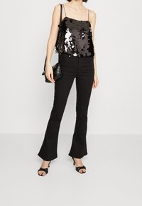 Błyszczący czarny tank top z cekinami na cienkich ramiączkach, zestawiony z czarnymi spodniami w dzwon i czarnymi sandałami na wysokim obcasie. Trzyma małą czarną torebkę.