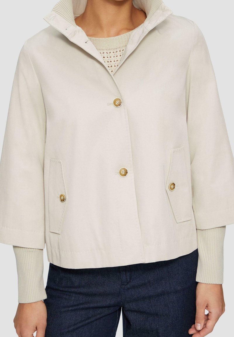 Beige cropped jasje met een staande kraag, twee vozakken en gouden knopen. De stof lijkt glad met een subtiele glans.