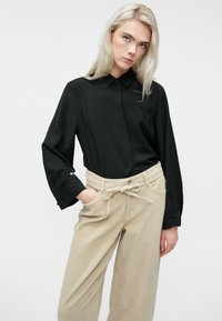 Jauna moteris su ilgais šviesiais plaukais, vilkinti juodą marškinį su mygtukais ir šviesiai rudos spalvos plačiomis šonkaulinėmis kelnėmis su surištu diržu.