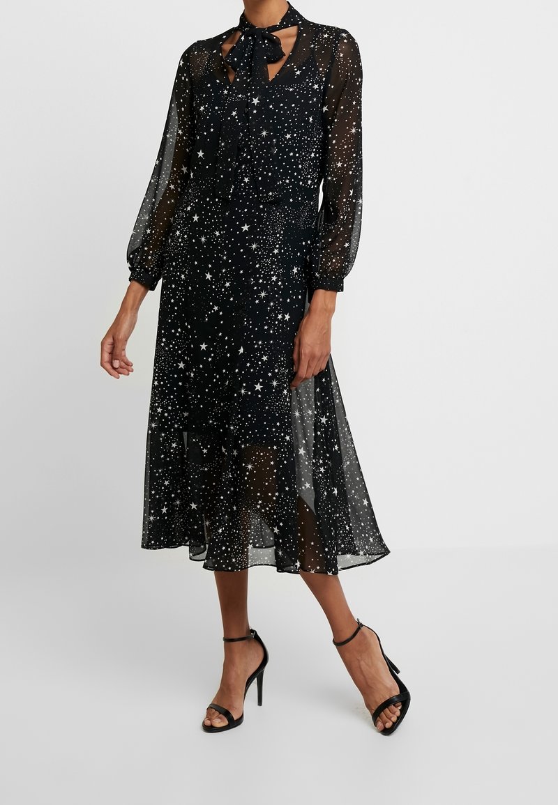 Femme portant une robe midi noire transparente avec motif étoiles blanches, manches longues, détail nœud au cou et sandales noires à talons.
