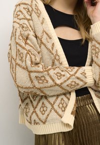 Stickad kofta i krämfärgad med brunt blommigt diamantmönster, med ribbat nederkant och ärmslut. Bärs över en svart ribbad topp.