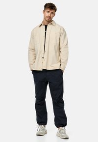 Beige knapjakke med to brystlommer, parret med mørkeblå bukser og lysegrå sneakers. Enkel design og afslappet pasform.