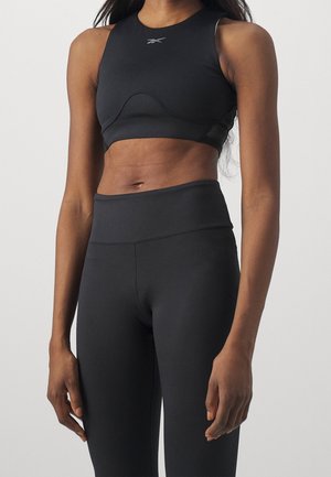 Atletische vrouw draagt een zwart mouwloos crop top en zwarte legging met hoge taille, staand tegen een effen achtergrond.