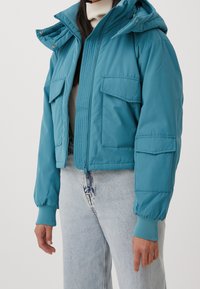 Szara krótka kurtka z gładkiego materiału, z zapięciem na zamek, dwiema kieszeniami na piersi i podszytym kapturem, w zestawie z jasnoniebieskimi dżinsami.