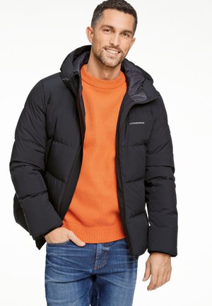 Mann trägt schwarze Steppjacke über leuchtend orangefarbenem Pullover und blauen Jeans, lächelt mit einer Hand in der Tasche vor weißem Hintergrund.