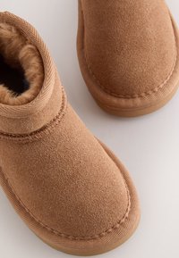 Bottes chelsea en daim marron avec une doublure douce en fourrure, bout arrondi et semelle en caoutchouc texturée. Détails de couture autour des bords.
