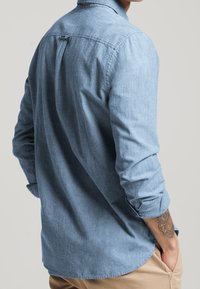 Lichtblauw overhemd in denimstijl met lange mouwen, knoopmanchetten en een lusdetail in het midden van de achterkant. De stof heeft een licht gestructureerde afwerking.