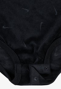 Μαύρο κορμάκι με απαλή υφή, διακοσμημένο με διακριτικά σκοτεινά λογότυπα Nike. Κούμπωμα με κουμπιά πίσω στο κάτω μέρος, στρογγυλεμένες άκρες, ελαστικές ταινίες στους ιμάντες.