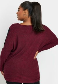 Maglione oversize a maniche lunghe in un ricco color bordeaux scuro, caratterizzato da una texture morbida, spalle scese e pannelli laterali a contrasto neri.