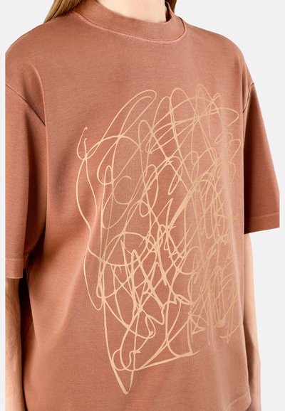Lyhythihainen T-paita pölyruusuisena, valmistettu pehmeästä materiaalista. Edessä on abstrakti beige graafinen kuvio. Pyöreä kaula-aukko.