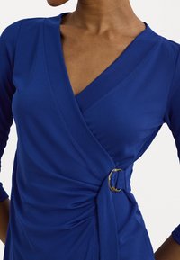 Blue wrap dress with a V-neck and three-quarter sleeves. Features a gold ring at the waist, textured fabric, and draped detailing.