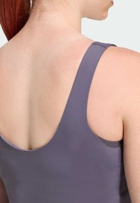 Nahaufnahme des oberen Rückens und der Schulter einer Person, die ein ärmelloses lila Oberteil trägt und rote Haare nach hinten gebunden hat.