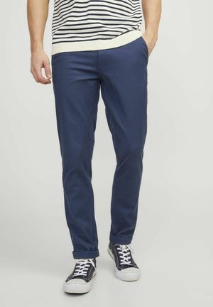 Chino in cotone blu navy con vestibilità slim, dotati di tasche laterali e risvolti. Indossati con sneakers stringate bianche e nere.