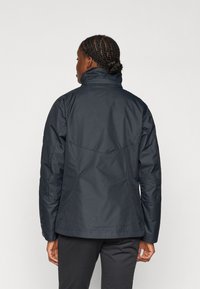 Marineblauwe, lichte jas met een hoge kraag, met een slank ontwerp en een gestructureerde stof. De jas eindigt op heuphoogte met een iets getailleerde pasvorm.