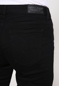 Musta denim-farkut, joissa on vartalonmyötäinen malli, takataskussa kaareva kirjailudetaili ja brändätty etiketti vyötäröllä.